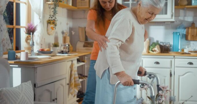 Satisfied, happy 90s old grandmother has support and assistance from her adult daughter or life assistant at home, uses a walker in the kitchen, and gets help to sit in armchair.