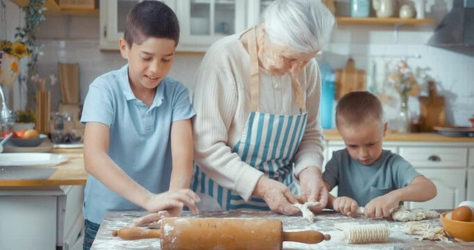 A multi-generational happy family with children enjoys cooking together in the kitchen. 90s grandmother teaches her grandsons how to knead dough using a kitchen tool while preparing a pie.