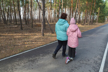Little girl riding scooter while walking with her grandmother in the forest on a sunny day
