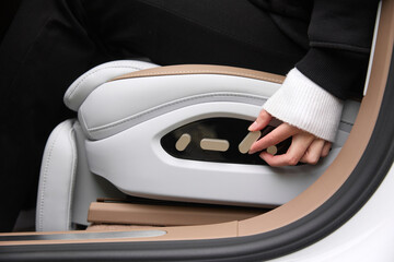 A close-up of a person's hand interacting with the controls of a modern, luxurious car seat. The image showcases a sleek and minimalist design featuring white and beige leather upholstery with precise