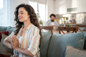 Serene woman meditating on sofa while man works on laptop in background