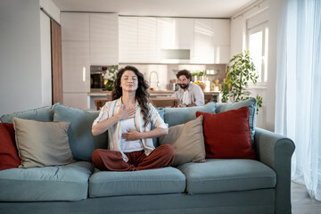 Woman practicing meditation on sofa while man working in background