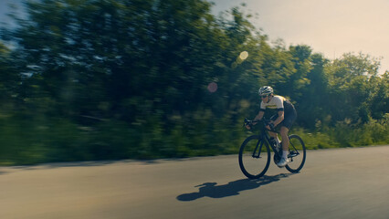 Obraz premium A helmeted cyclist rides down a scenic road, surrounded by lush greenery. The motion blur effect emphasizes speed and intensity. Aerial view.