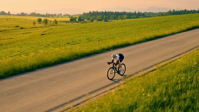 A person in a helmet rides a bicycle on a rural road, surrounded by lush green fields. The aerial view showcases the cyclist's endurance and the serene countryside landscape.