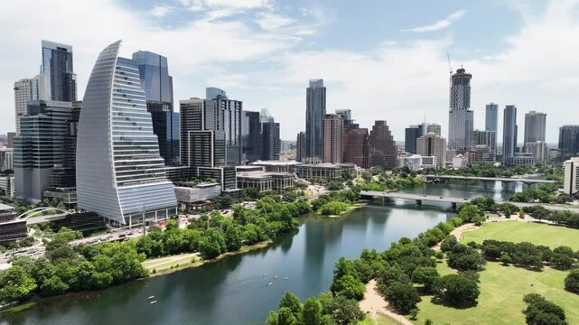 Austin's Lady Bird Lake by day. Lush green trees, lawn. Downtown skyline view. Kayaks, running people. Active traffic.