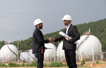 Two Business Man or Investor with black suit and safety hat working with drawing and digital tablet due business at front Refinery and gas petrochemical plant  