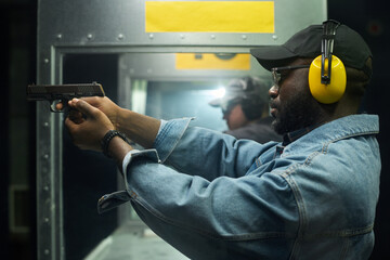 Man wearing headphones aiming pistol at indoor shooting range with focus and precision, another...