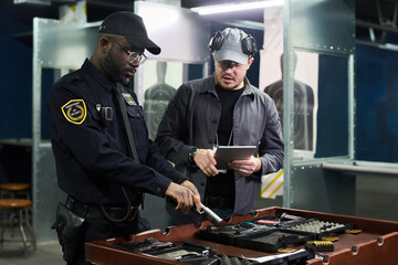 Two men in uniform collaborating in shooting range, examining equipment and taking notes on tablet....