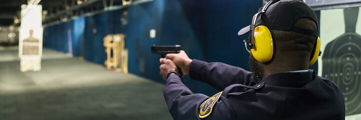 Man aiming at target in indoor shooting range, wearing safety gear and ear protection. Environment includes visible targets and safety barriers in controlled setting