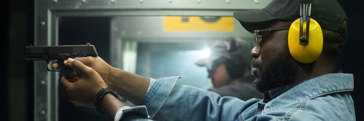 African American man with safety gear, including earmuffs and eyewear, aiming handgun at target in indoor shooting range while instructor observes in background © pressmaster