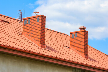 Red tiled roof with two prominent chimneys and gutters, set against a bright blue sky with clouds,