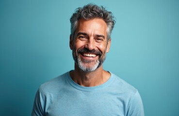 Obraz premium Handsome middle-aged man smiles at camera against blue background. Cheerful male with gray hair and beard looking happy. Casual portrait. Smiling, positive, adult person in studio.