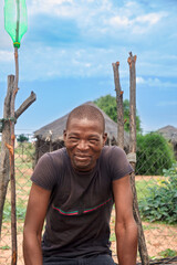 african village single smiling young african man, sitting in front of mud house with thatched roof, rural living