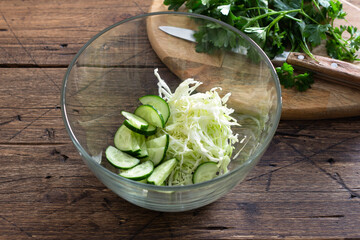 Fresh cucumber and cabbage salad in glass bowl on wooden table, healthy vegetarian food preparation concept
