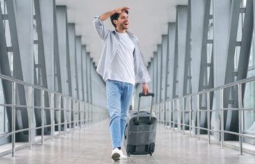 Looking forward to new places. Cheerful man walking with luggage at airport, ready for boarding on...