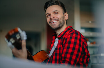 Smiling young man in red plaid shirt playing acoustic guitar at home. Relaxed indoor scene with...