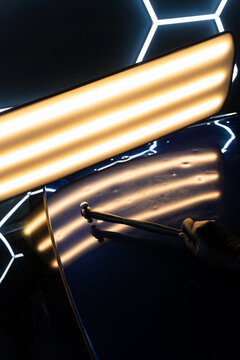 PDR technician holds a specialized hammer against a reflection light board to check dent correction on the hood. Accurate paintless car dent removal.