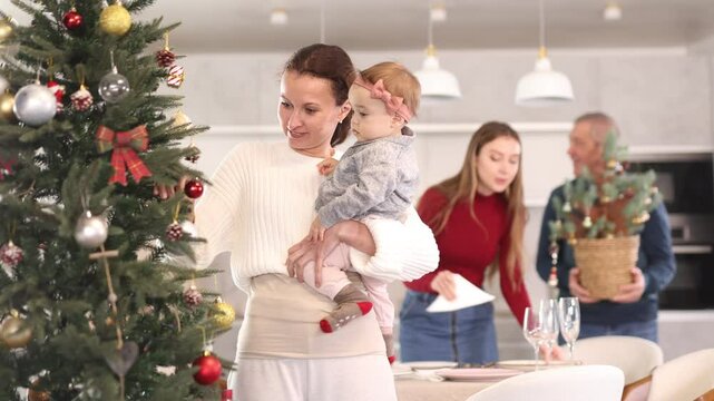 Young woman with a baby in her arms decorates a Christmas tree with toys while the rest of the family prepares a festive dinner. High quality 4k footage
