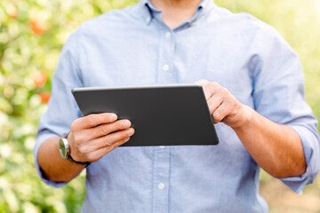 Smart farm management, growing organic fruits, control cultivation with mobile device. Millennial guy working on digital tablet at garden, on green leaves background, cropped, close up, copy space © Prostock-studio