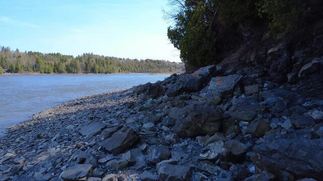 Low‑altitude drone footage of a rocky, shaded riverbank along the Mitis River under a clear blue sky in Quebec, Canada, 2025.