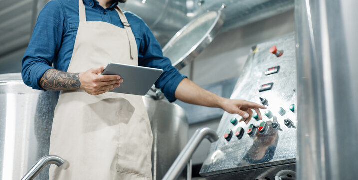 Male worker on beer factory, management and mobile app. Busy male employee or owner in apron with digital tablet controls equipment at brewery and push button, cropped, free space, panorama