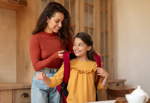 Young Arabic Mom Putting Backpack On Daughter's Shoulders Sending Her To School, Preparing Preteen Schoolgirl Kid For Classes At Modern Home Interior. Morning Preparation, Schooler Gets Ready