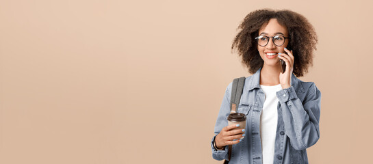 Modern worker, freelancer or blogger. Smiling teen african american curly lady in glasses with backpack, speaks on phone and holds cup of coffee takeaway, isolated on light background, free space