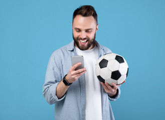 Happy young guy with soccer ball using smartphone, winning sports bet, rooting for his favorite team on blue studio background. Millennial man watching online football championship