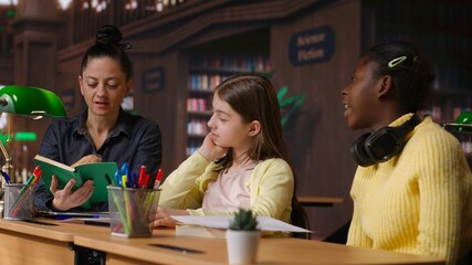 Female tutor mentoring schoolgirls in a school library setting, boosting their understanding of a subject through personalized guidance. Teacher doing private lessons with two pupils. Camera B.