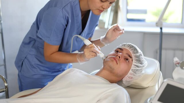 Young female cosmetologist performs hardware facial microcurrent procedure to young male patient