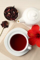Delicious hibiscus tea in cup, teapot, dry roselle sepals and flower on light wooden table, flat lay