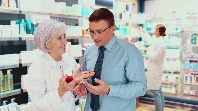 Interested middle-aged man asking old female apothecary for advice concerning preparation in box in drugstore 