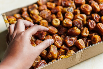 A hand reaches into a box filled with soft dates. The textured skin of the dates are a vibrant, rich brown. This image creates a sense of healthy, wholesome and organic food.