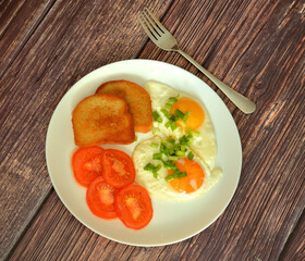 Two fried eggs sprinkled with green onions, tomato slices and wheat croutons on a wooden table.