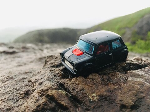 A black classic Mini Cooper toy car perched on a rugged, wet rock surface with a misty, green hillside in the background.
