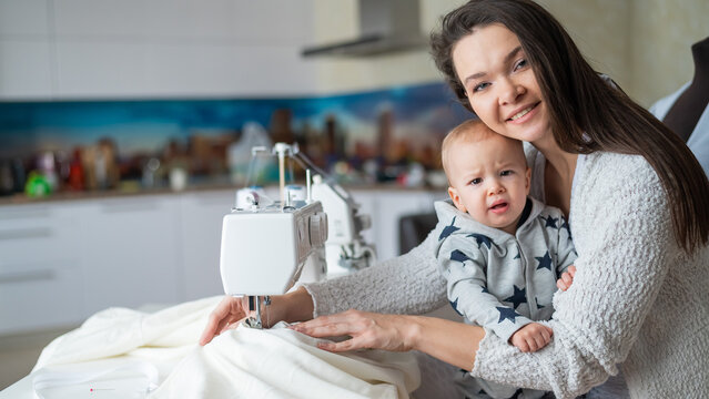 A young woman sews in the kitchen and holds a small child. Mom teaches her little son to sew on a sewing machine. Self-isolation homework