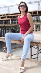 Fototapeta premium Stylish young woman in a red top and blue jeans sits confidently on a wooden bench on outdoor stands, showcasing a modern, casual look with platform sandals