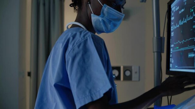 A healthcare professional in scrubs and mask checks a monitor in a dimly lit room. The side angle adds a documentary video feel.