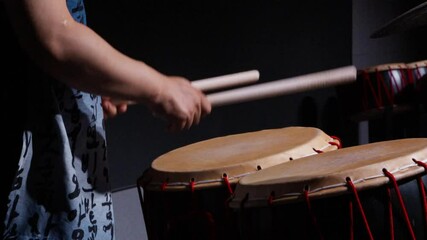 Close-up of hands striking a traditional Korean buk drum with wooden sticks, showing drumhead and red lacing detail. Close-up shot