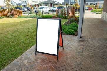 Blank white A-frame sandwich board mockup on a brick-patterned patio outside a suburban commercial...