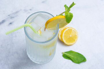 A glass of refreshing lemon water with ice cubes, mint leaves and a straw stands on a light marble...