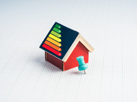 House energy efficiency rating class, cost savings and low-energy eco-friendly home concept. Auditor rate label on roof of red miniature house and blue thumbtack pin on white graph paper background.