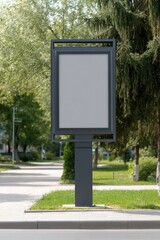 Blank rectangular billboard on a dark grey pole stands on a paved sidewalk beside a grassy area, flanked by trees