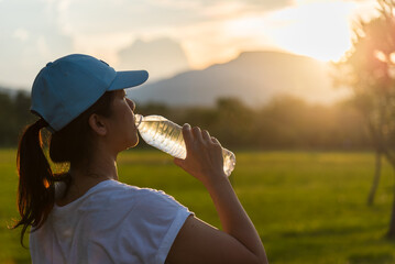 Asian woman drinking water after running and training outdoors at sunset. Healthy girl drinking water from bottle for outdoor exercise, running workout or fitness training concept.