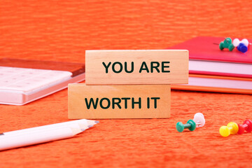 YOU ARE WORTH IT words made of wooden blocks on an orange background with stationery lying next to...