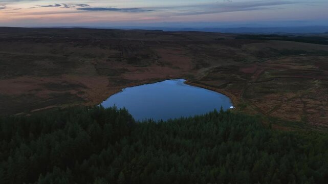 Bragan, County Monaghan, Ireland, September 2022. Drone orbit clockwise over dense forest along edge of Lake Menish establishing surrounding bog land at blue hour dusk.