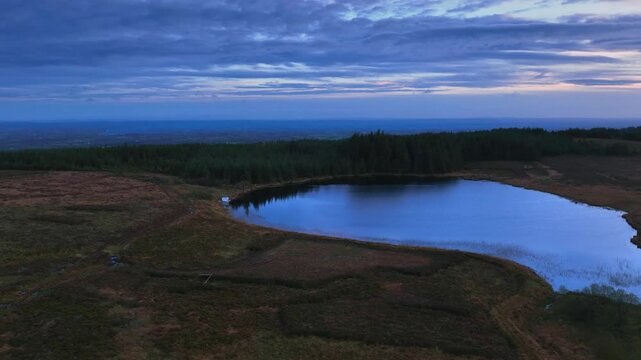 Bragan, County Monaghan, Ireland, September 2022. Drone establishing low orbit clockwise around Lake Menish with surrounding bog wetlands at blue hour with purple pink haze and yellow glow in sky.