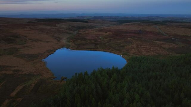 Bragan, County Monaghan, Ireland, September 2022. Drone orbit medium establishing clockwise above Lake Menish with soft purple haze glow in sky and reflection in water in serene bog land fields.