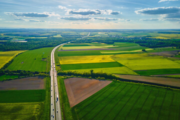 Aerial view of highway cutting through agricultural fields with blooming rapeseed crops. Transportation infrastructure among natural farmland. Spring countryside landscape