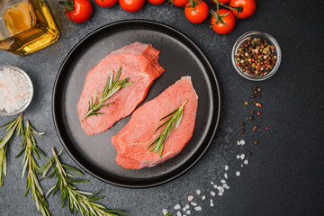 Raw veal steaks on black ceramic plate with rosemary cherry tomatoes salt oil and pepper